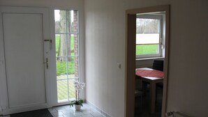 Una habitación con una puerta blanca, un ventanal con vista a un muro de ladrillo y follaje, y una mesa con mantel rojo.