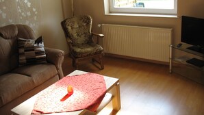 Una sala de estar con sofá, un sillón floral, una mesa de centro con un paño rojo, televisor sobre un mueble bajo y ventana con vista a follaje.