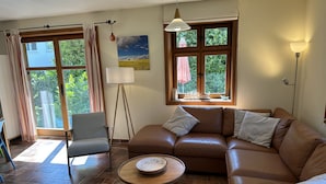 Un salon avec un canapé en cuir marron, une table basse ronde en bois, un lampadaire et une vue sur la verdure grâce à de grandes fenêtres.