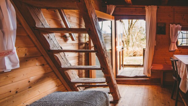 Interior de una cabaña de madera con techo inclinado, escalera de madera, una mesa con sillas y una ventana con cortinas.