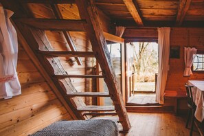 Interior de una cabaña de madera con techo inclinado, escalera de madera, una mesa con sillas y una ventana con cortinas.