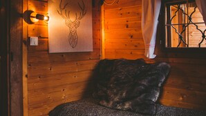 Una habitación de cabaña acogedora con interior de madera, una cama con manta oscura, una lámpara fijada a la pared y una ventana con cortinas.
