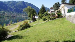 A lakeside view with a grassy slope, a white building with a green fence, and mountains in the background. - Directly on the lake with terrace (Tre Capitelli)