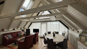A spacious loft with a high ceiling, exposed wooden beams, a fireplace, a dining table with chairs, a sofa, and a TV. - With sauna and fireplace (Rheinboellen)