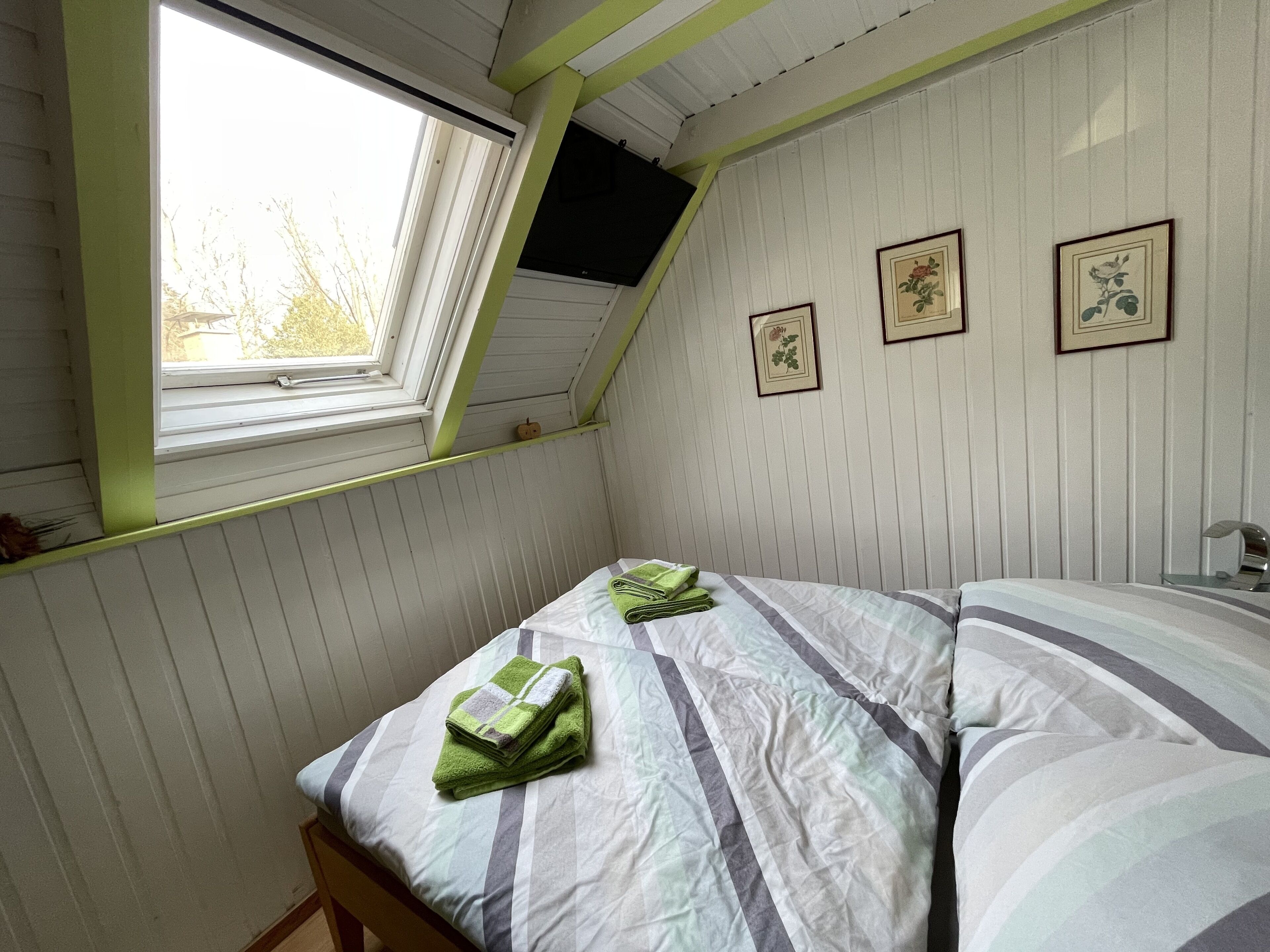 A room with a sloped ceiling, a bed with striped bedding, a window, and framed pictures on the wall.