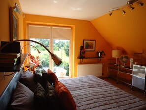 Un dormitorio con cama, escritorio con lámpara, radiador y ventana con vistas a zonas verdes.