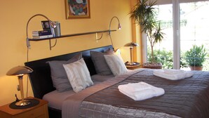 Ein ordentlich bezogenes Bett mit einer grauen Tagesdecke, eine Nachttischlampe, ein Regal mit Büchern und ein Bild an der Wand.