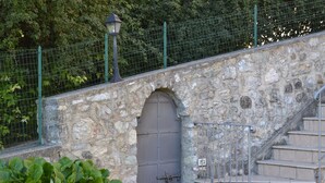 Un escalier en pierre mène à un portail en métal, qui est lui-même entouré d’une clôture verte et d’une végétation luxuriante.
