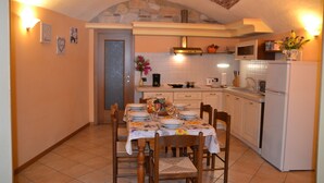 On aperçoit un coin repas avec une table dressée pour quatre personnes, entourée de chaises en bois. Au fond, on distingue une cuisine avec des appareils électroménagers blancs et une arche en pierre.