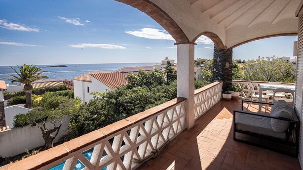 A balcony with a view of the sea, a pool, and a seating area with a table and chairs.