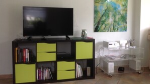 A living room with a flat-screen TV on a black stand, a bookshelf with green and black shelves, a painting of trees, and a white desk with a computer and a printer. - Only 250m to the beach - for 4 pers., 70sqm, large balcony. South-facing garden/terrace, conservatory (Schönberger Strand)