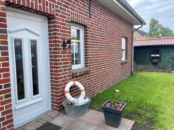 A brick house with a white door, a window with a curtain, and a metal bucket with a life preserver.