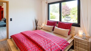 Une chambre à coucher avec un lit, une fenêtre avec des rideaux, une table de chevet avec une lampe, et une vue sur la verdure à l’extérieur.