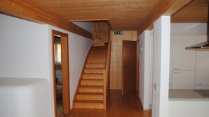 Un escalier en bois menant à un deuxième étage, un coin cuisine avec des meubles blancs et une porte portant une indication de sortie.
