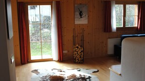 Une pièce aux murs en bois, avec une grande fenêtre, un tableau sur le thème du ski et un tapis en peau de vache sur le sol.