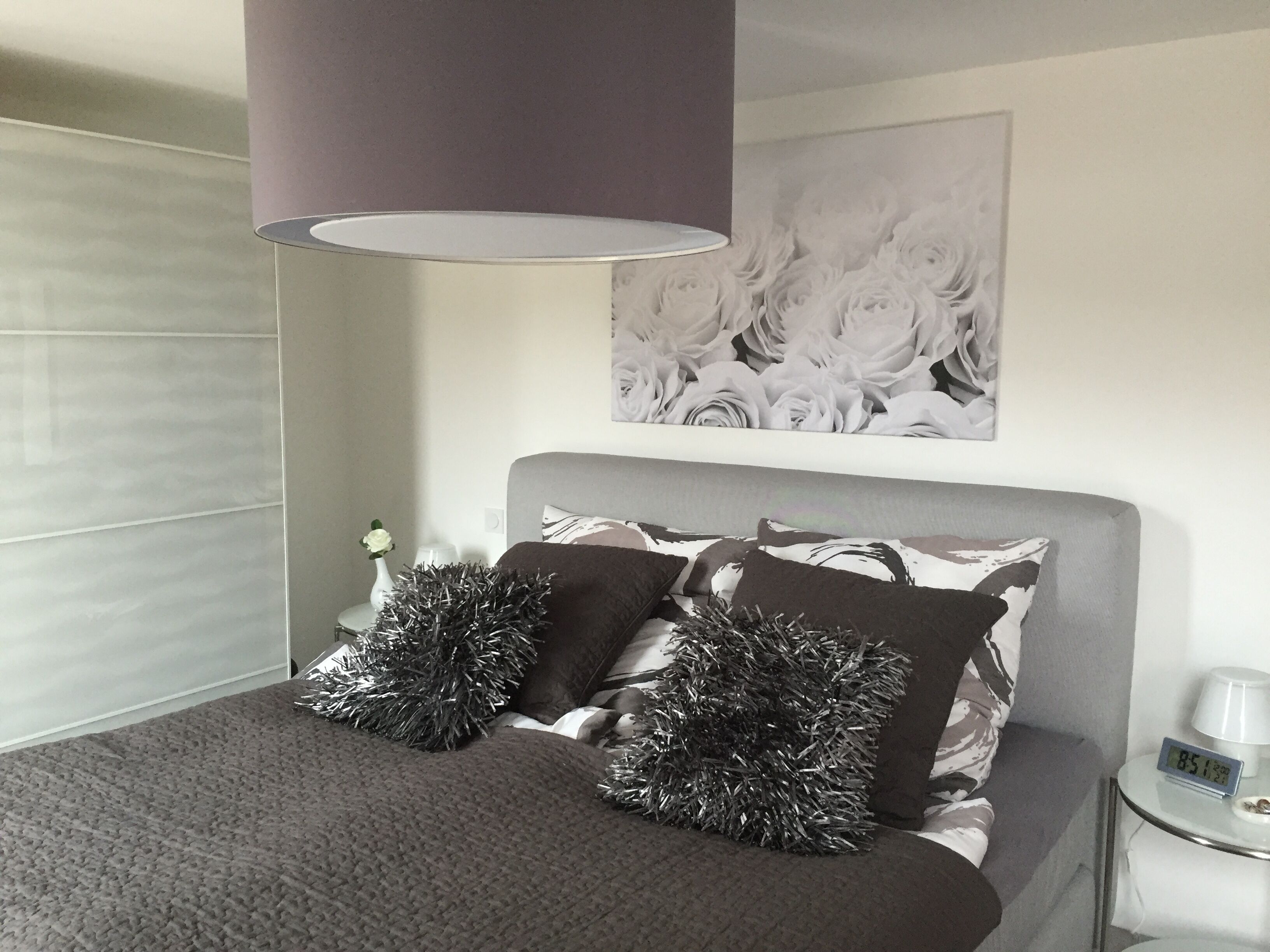 A modern bedroom with a grey headboard, two decorative pillows, a bedside table with a clock, and a wall-mounted artwork.