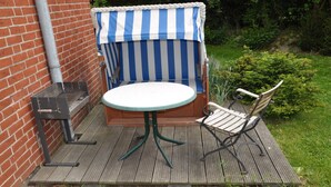 Una terraza de madera con una mesa redonda y dos sillas, un toldo a rayas y una pared de ladrillo.
