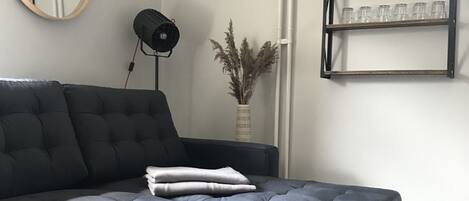 A modern living room with a dark grey sectional sofa, a round coffee table, a wall-mounted mirror, a floor lamp, a vase with dried plants, and a shelf with glasses.