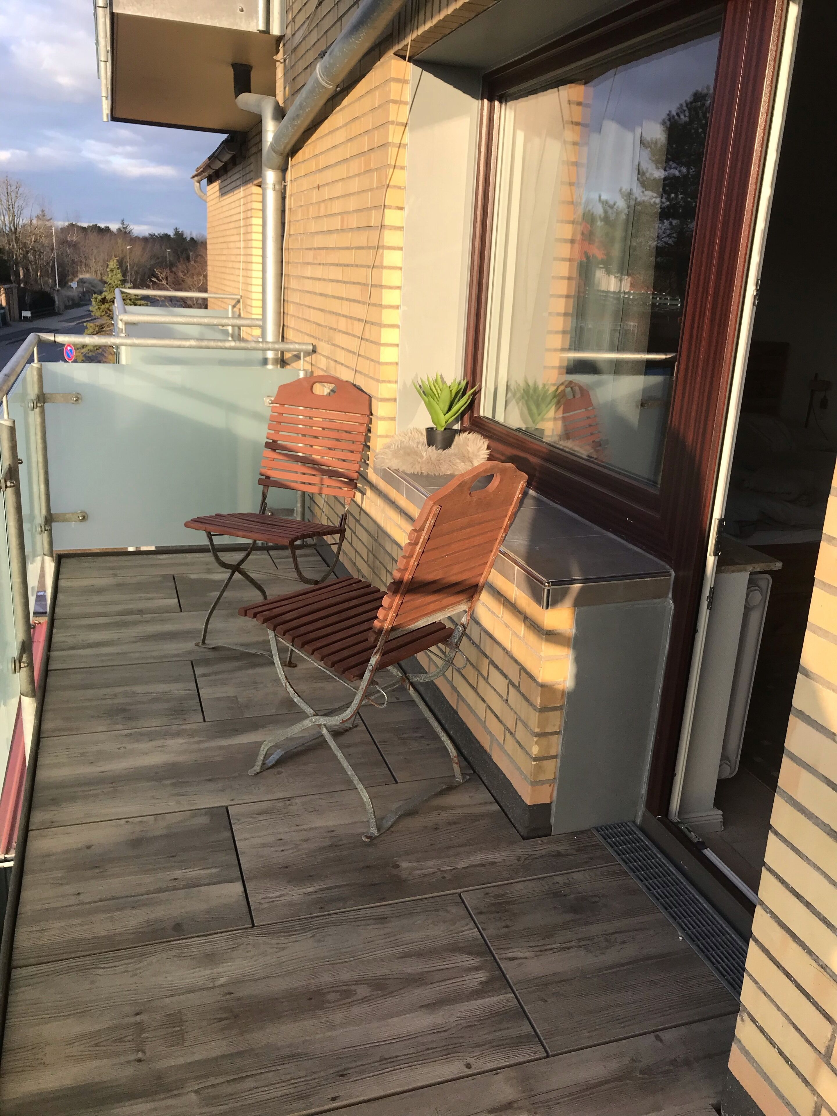 A wooden deck with two chairs and a small table, a potted plant, and a window with a view of trees.