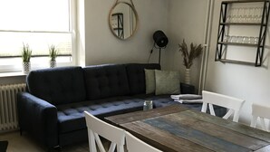 Un comedor con una mesa de madera, sillas blancas, un sofá oscuro y una mesita auxiliar con una lámpara.
