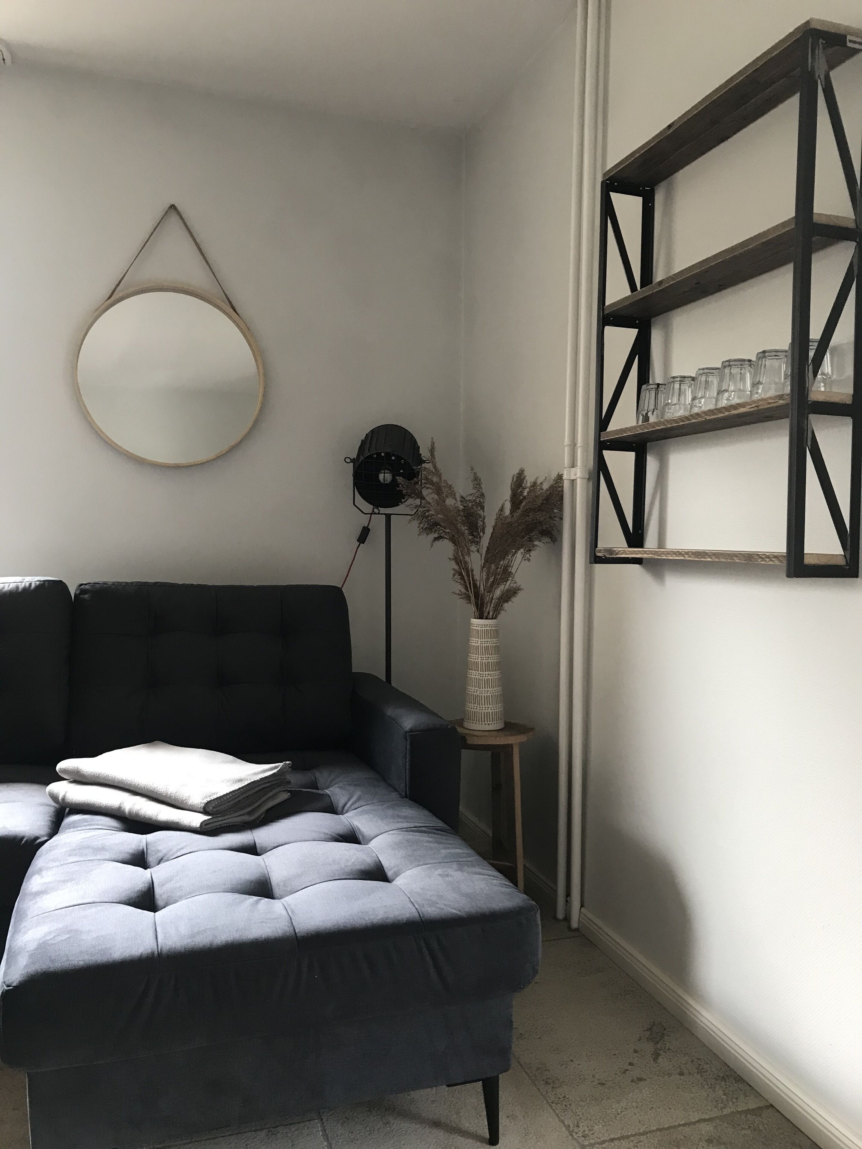 A modern living room with a dark grey sectional sofa, a round mirror, a floor lamp, a small wooden table with a vase, and a black metal shelving unit with glasses.