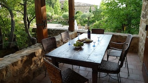 An outdoor patio with a wooden table set for dining, surrounded by wicker chairs, and a stone wall.