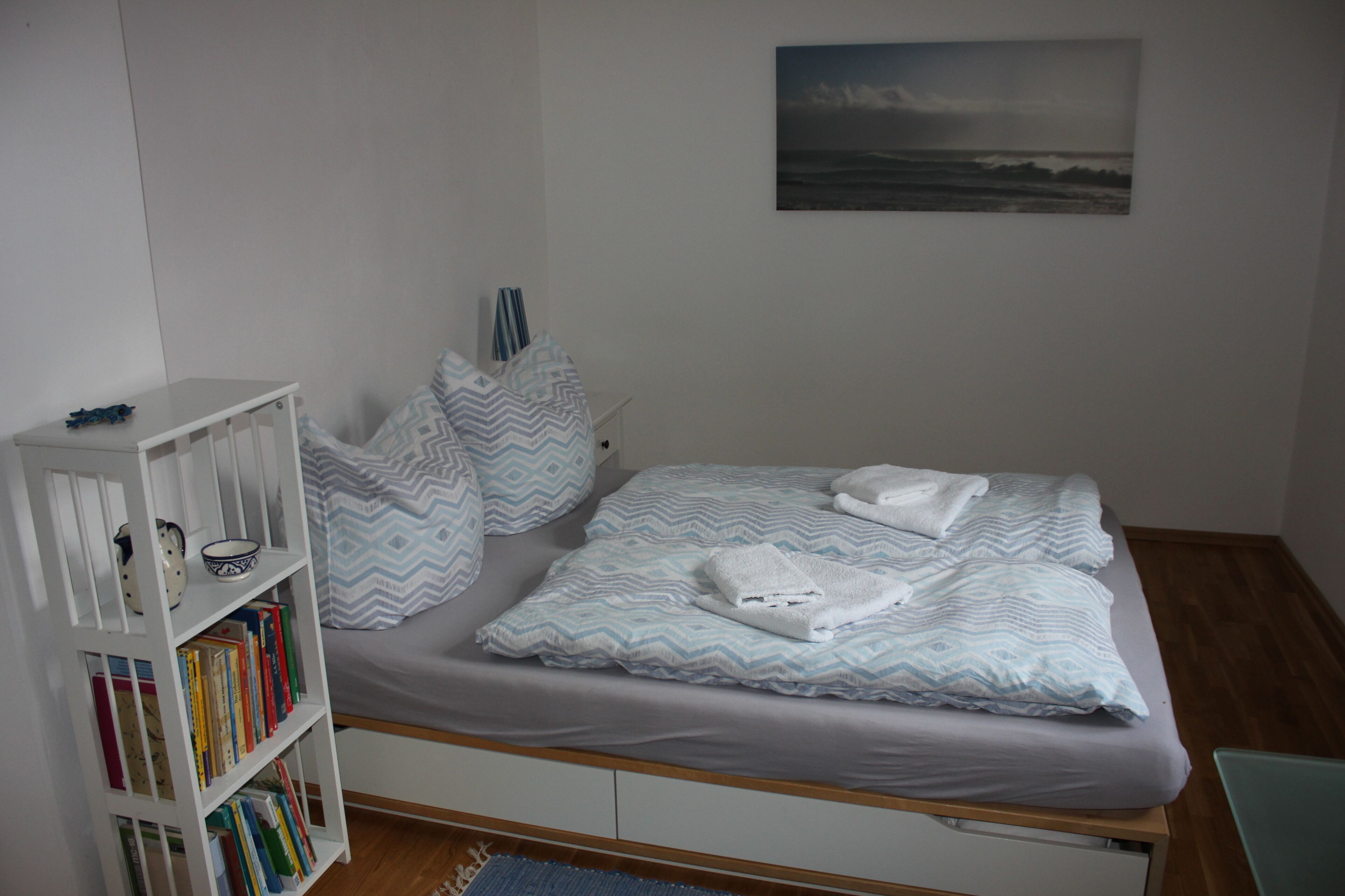 Ein Schlafzimmer mit Bett, Kissen, einem Bücherregal und einem Bild an der Wand.
