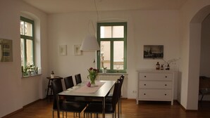 Un coin repas comprenant une table, des chaises, une fenêtre avec des volets verts, un radiateur et un meuble contenant des bouteilles.