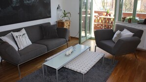 Un salon moderne avec un canapé gris, un fauteuil assorti, une table basse en verre et une vue sur l’extérieur grâce à une porte-fenêtre coulissante.