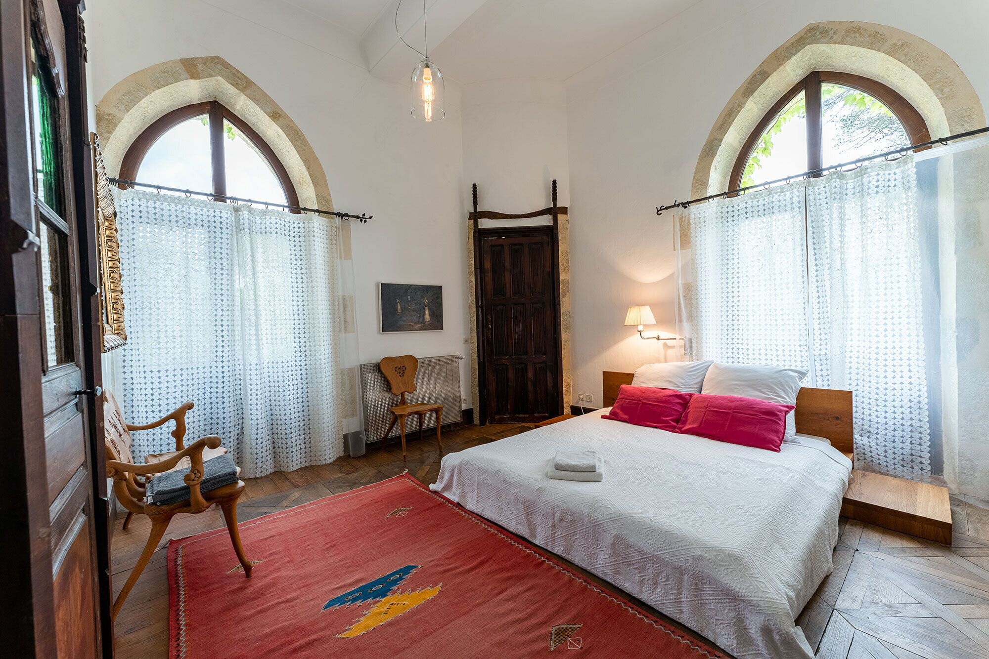 A bedroom with a bed, a chair, a wooden headboard, a red rug, and a wooden door.