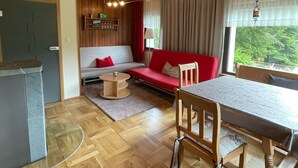 A modern living room with a wooden floor, a red sofa, a dining table with chairs, and a kitchen area with a fridge and shelves. - Detached vacation home with sauna and whirlpool for individual use (Thüringen)