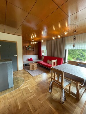 Ein modernes Wohnzimmer mit Holzboden, einem roten Sofa, einem Esstisch mit Stühlen und einer Küchenzeile mit Kühlschrank und Regalen.