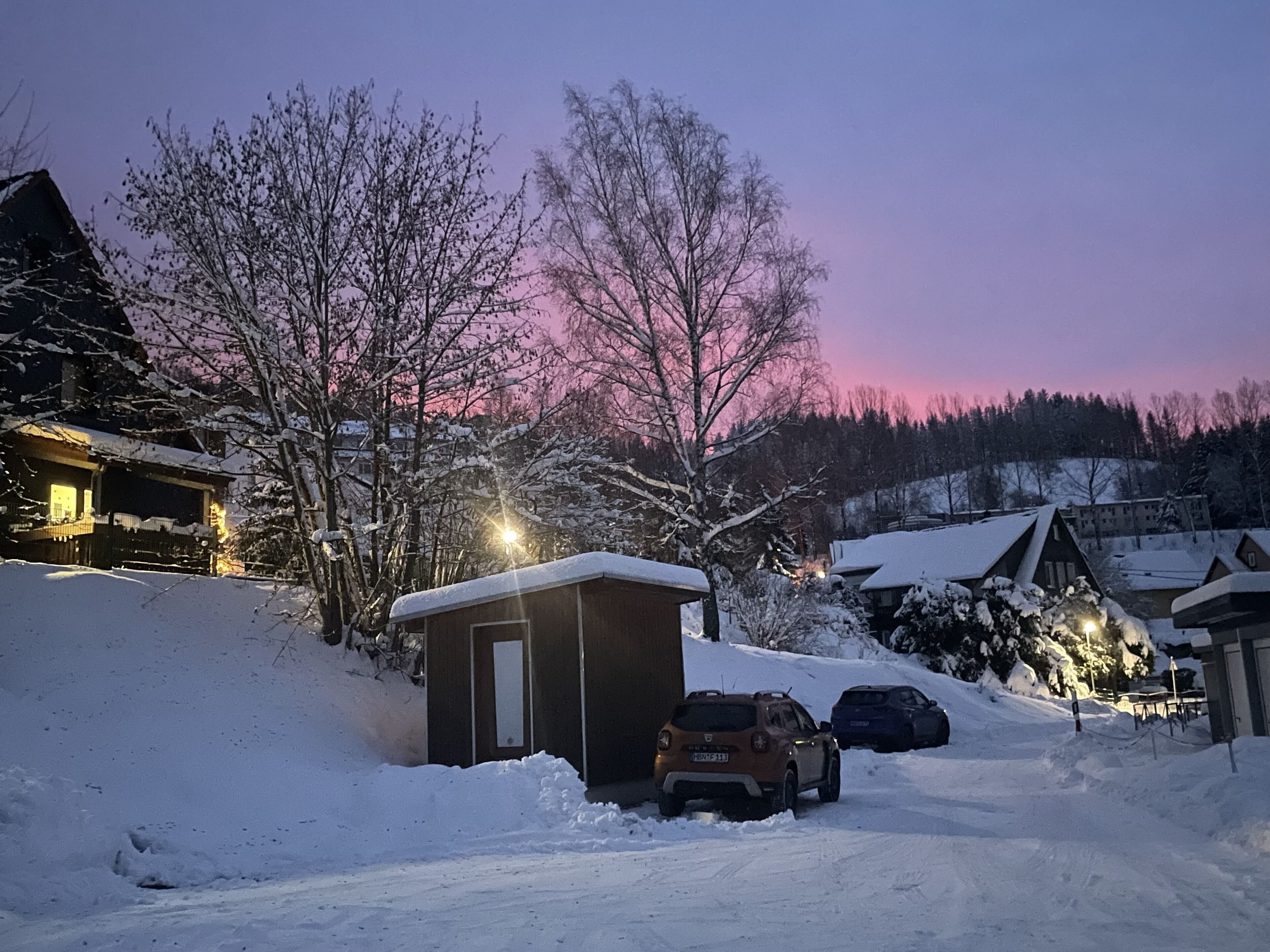Un quartier résidentiel enneigé, avec des maisons, un petit abri de jardin et des voitures garées.