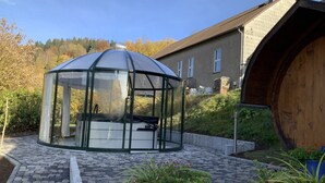 A glass-enclosed gazebo with a bench inside, surrounded by a stone-tiled patio and a wooden barrel. - Detached vacation home with sauna and whirlpool for individual use (Thüringen)