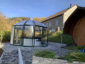 Ein überdachter Pavillon mit Sitzbank im Inneren, umgeben von einer Steinterrasse und einem Holzfass.