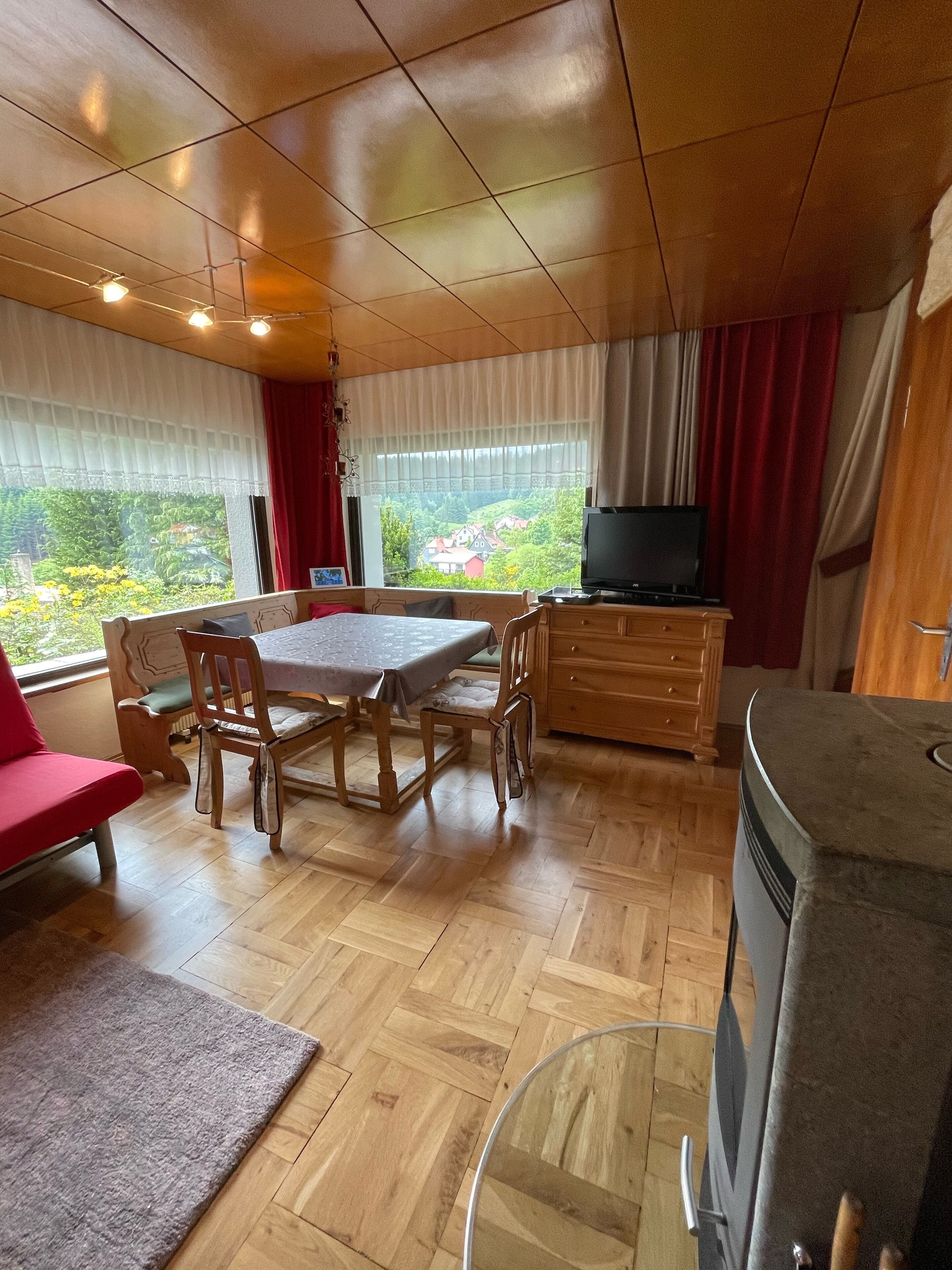 A room with a wooden table, chairs, a sofa, a TV, and a view of greenery outside.