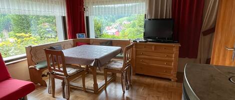 A room with a wooden table, chairs, a sofa, a TV, and a view of greenery outside.
