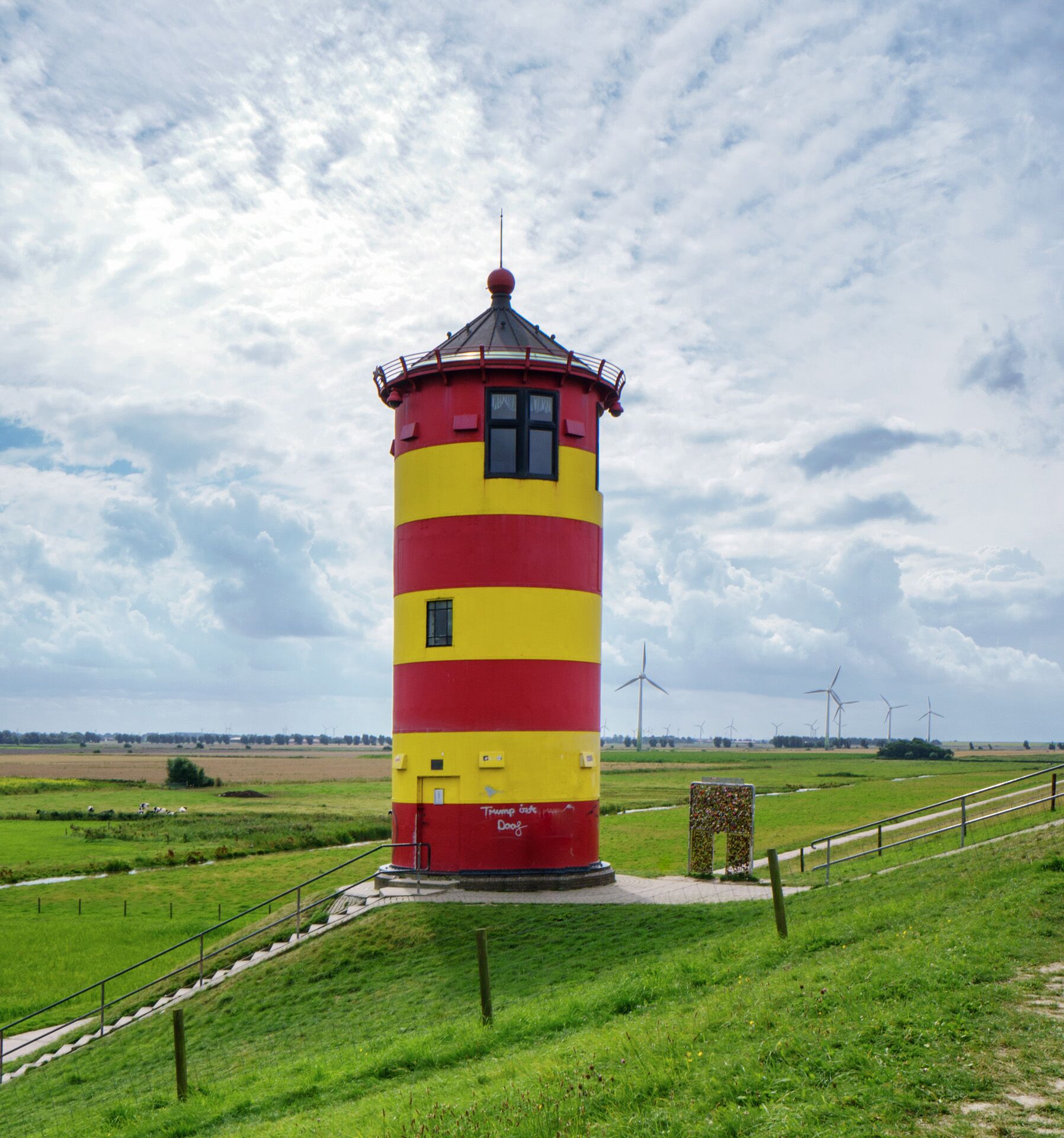 A cylindrical lighthouse with alternating red and yellow stripes, situated in a grassy field with wind turbines in the background.
