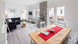 Un salon moderne avec une table à manger en bois, des chaises blanches, un canapé noir et un téléviseur fixé au mur.
