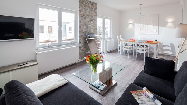 Un salon moderne avec une table basse en verre, des canapés gris, un téléviseur et un coin repas comprenant une table et des chaises blanches.