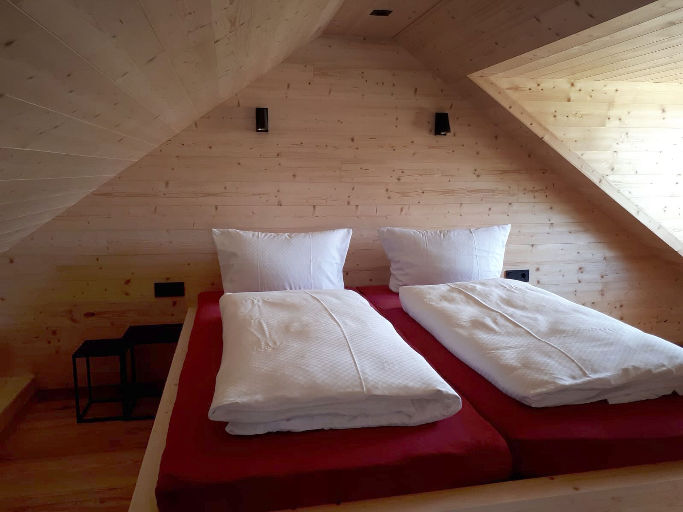 Habitació petita i acollidora amb sostre inclinat, parets de fusta i un llit amb roba de llit vermella.