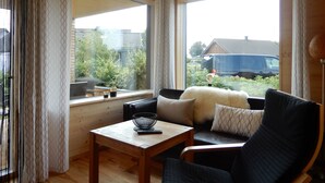 A cozy living room with a wooden ceiling, a large window with curtains, a sofa with cushions, a small table with a bowl and remote control, and a view of a garden and parked cars outside. - Rent a bungalow, 300 mtr to the beach, private ground, 4 pers. (Ostseeferiendorf Zierow)