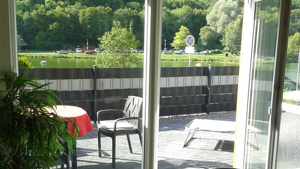 Une pièce dotée d’une porte-fenêtre coulissante donnant sur un patio extérieur aménagé avec une table et des chaises, un pot de fleurs et une vue sur un parc et un lac.