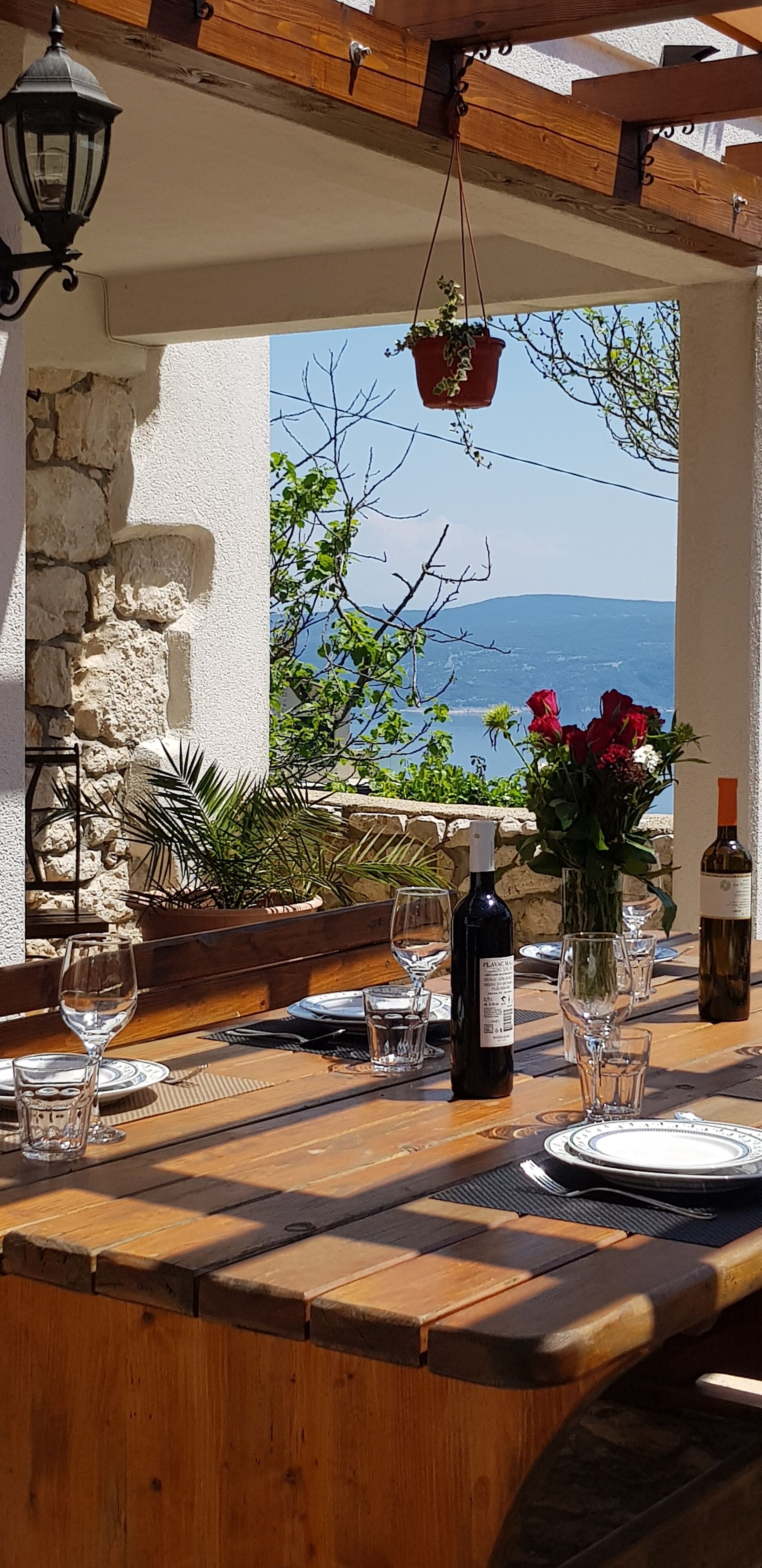 A wooden table set for dining with wine bottles and glasses, a hanging planter with greenery, and a view of the sea and mountains.