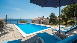 A swimming pool with lounge chairs and an umbrella, surrounded by a house and a view of the sea. - Vila Pavincica with swimming pool, soccer pitch and award-winning honey tasting (LABIN)