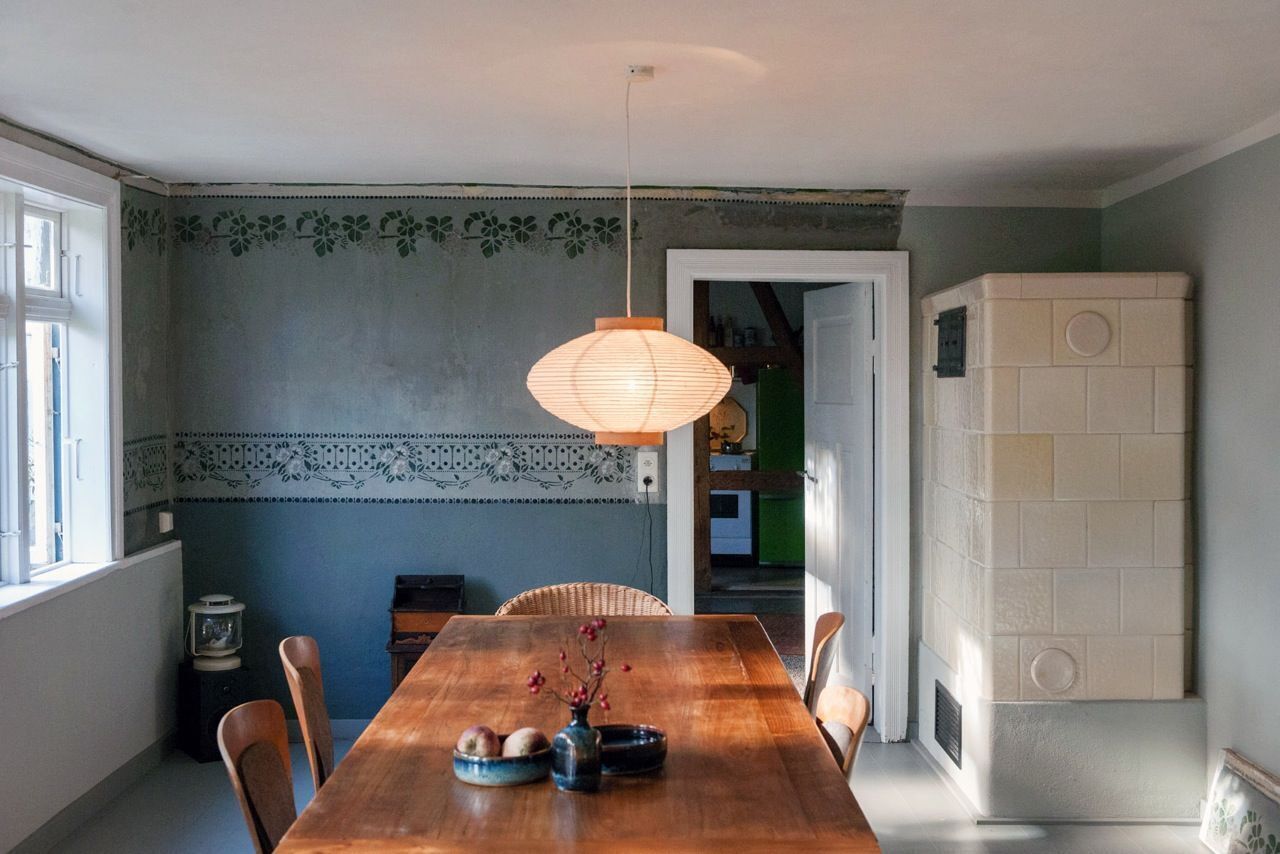 A dining area with a wooden table, chairs, a hanging light fixture, and a decorative wall with a floral pattern.