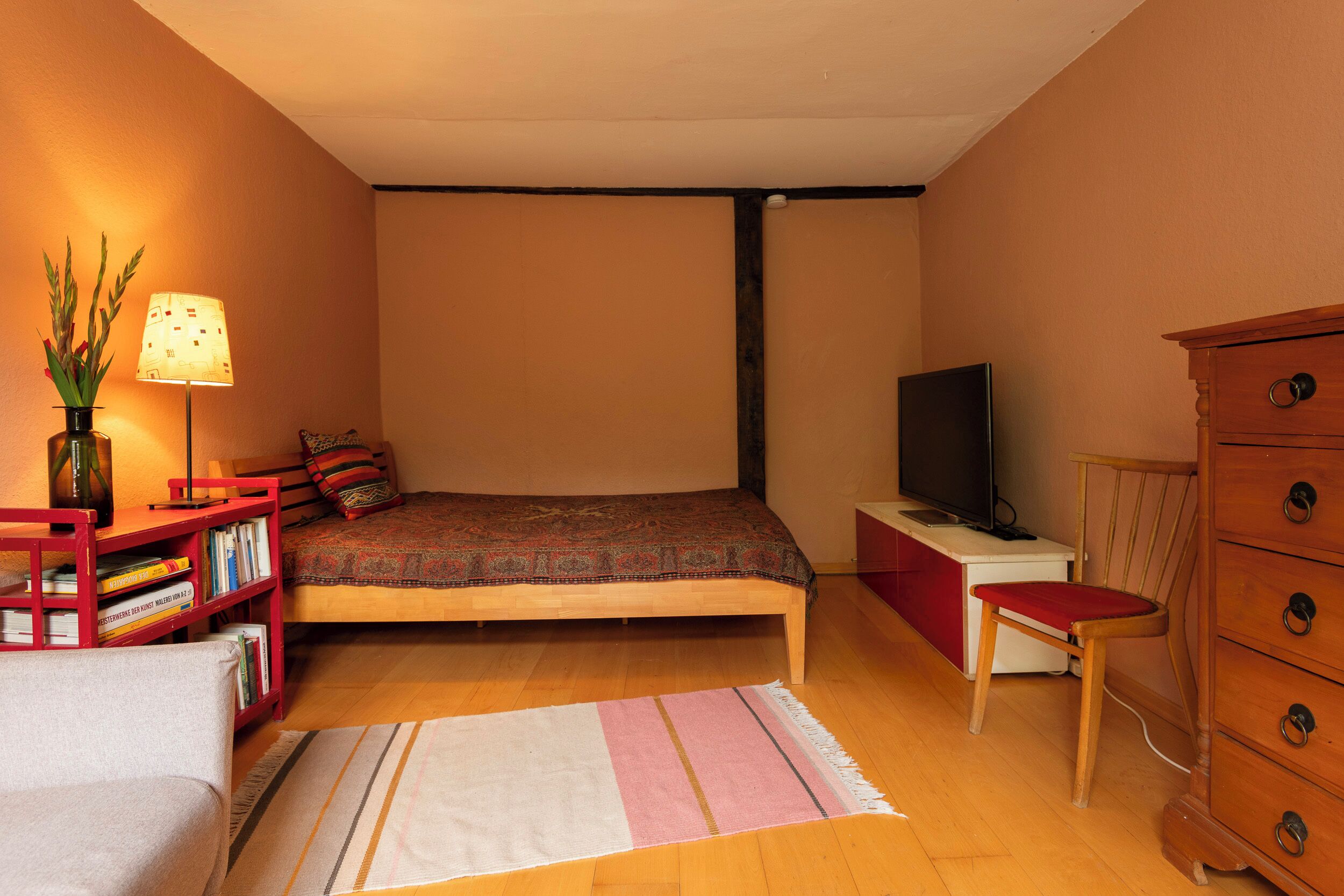 A bedroom with a bed, a television, a dresser, a chair, a bookshelf, and a lamp.