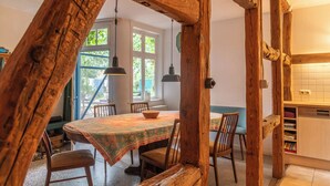 Uma sala de jantar com mesa e cadeiras de madeira, janela com vista para vegetação, e área de cozinha com armários e lava-louças.