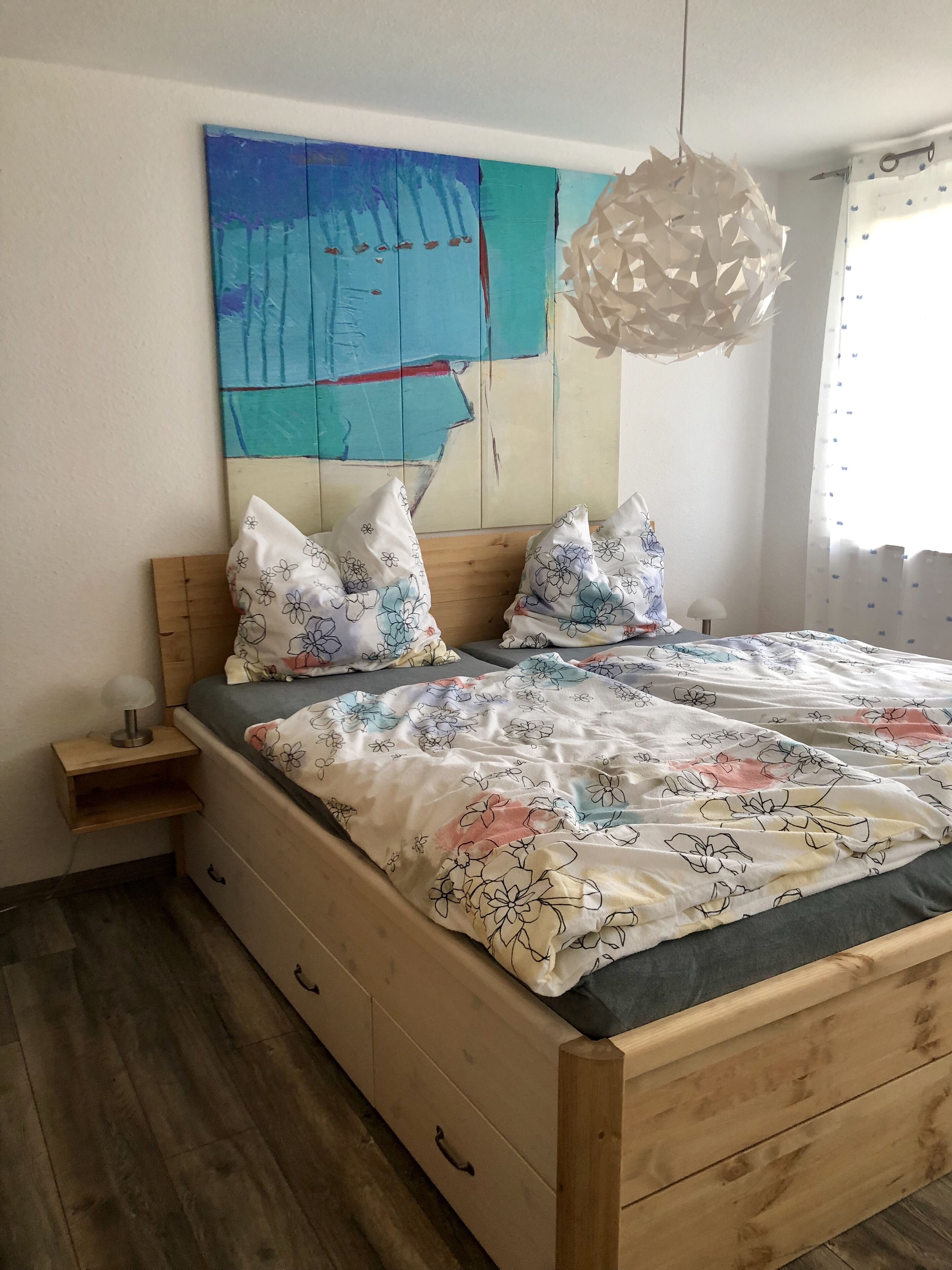 Une chambre à coucher avec un lit en bois, une literie à motifs floraux, une table de chevet, une lampe et un tableau abstrait aux couleurs vives accroché au mur.