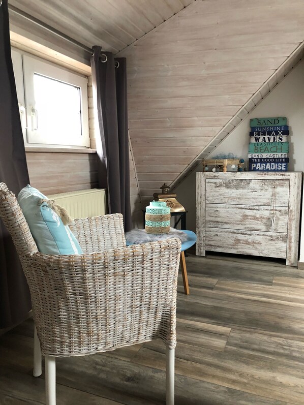 A room with a wicker chair, a small table, a decorative cabinet, and a window with curtains. - FH Meeresleuchten, 120 sqm, fireplace, sauna, coffee machine, dogs allowed. (Wangerland)